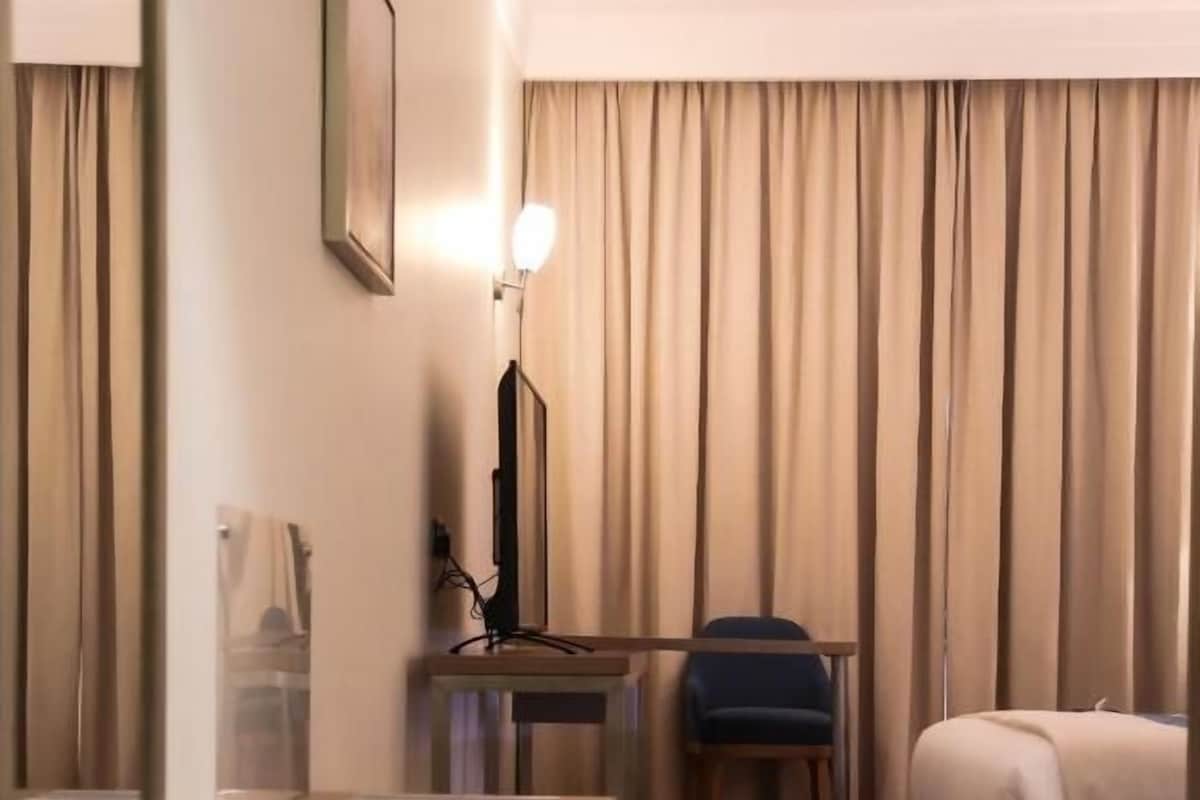 A hotel room with a bed, a desk with a chair, a lamp, and a large window with curtains.