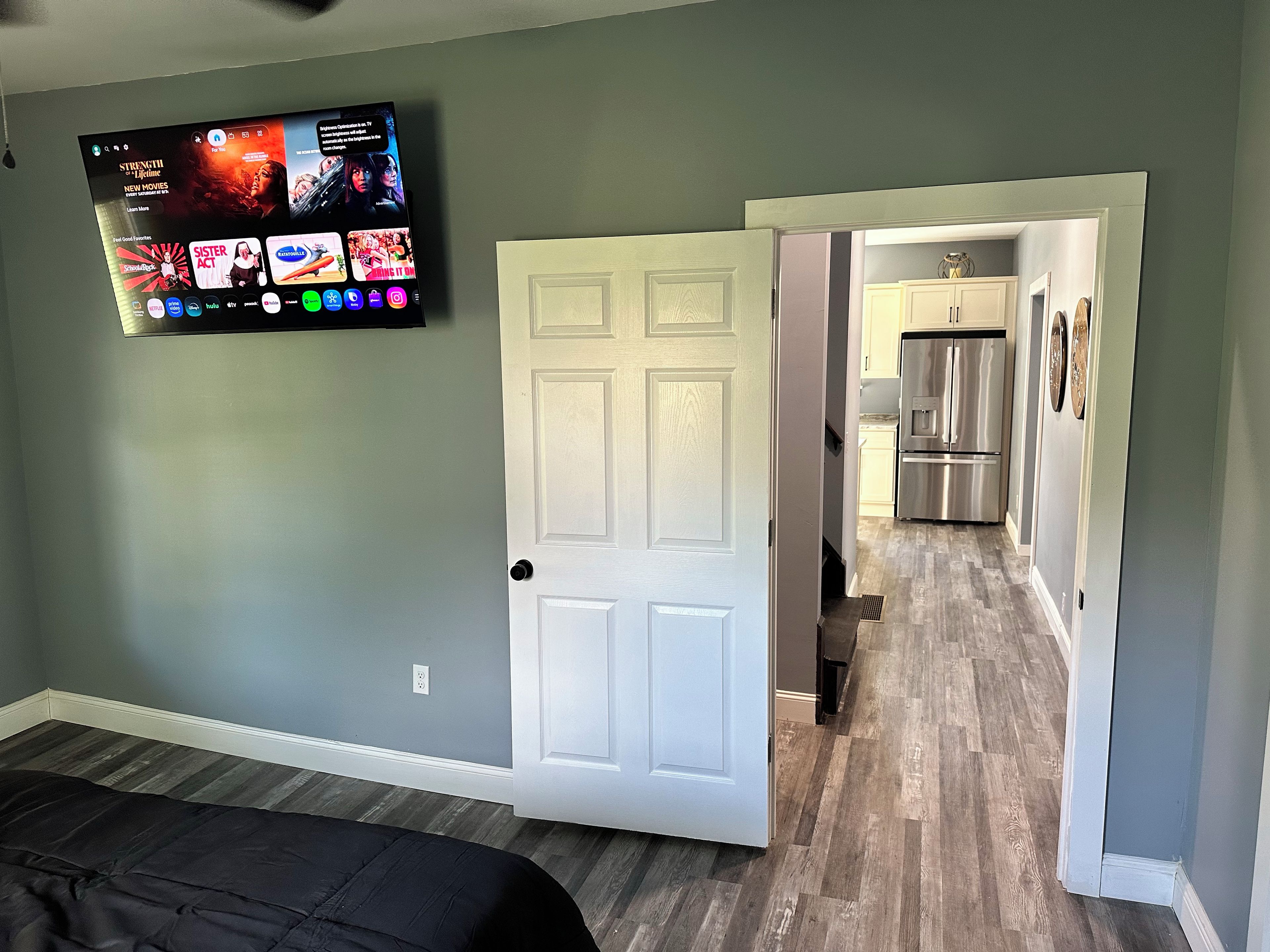 Une pièce comprenant un lit, un téléviseur fixé au mur et une porte donnant sur un espace cuisine.