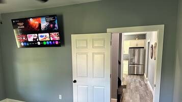 Une pièce comprenant un lit, un téléviseur fixé au mur et une porte donnant sur un espace cuisine.