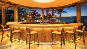 Een bar met een houten toonbank, barkrukken en flessen op planken.