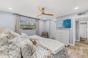 Ein Schlafzimmer mit Bett, Deckenventilator, Fenster mit Jalousien, einem an der Wand befestigten Fernseher und einer Tür zu einem Badezimmer.