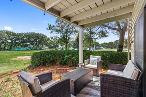 Une terrasse couverte, meublée de mobilier en osier, avec vue sur une pelouse bien entretenue et des arbres en arrière-plan.