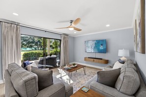 A modern living room with a ceiling fan, a flat-screen TV, and a large window with curtains. - The Sandpiper Pearl - Seascape Villa 2A *Updated*Quiet villa*Beach access* (Miramar Beach)