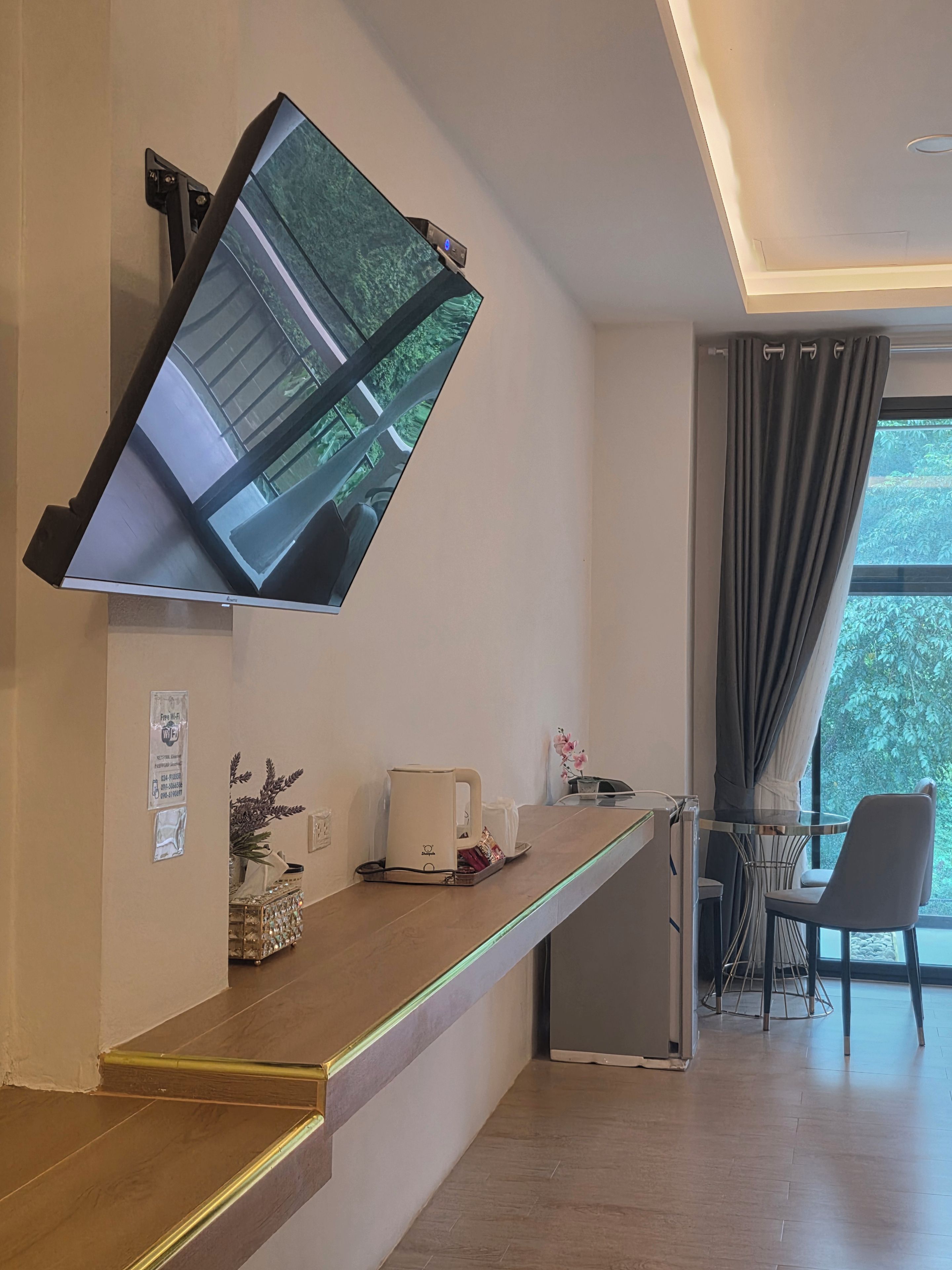Une chambre d’hôtel moderne dotée d’un téléviseur à écran plat fixé au mur, d’une petite kitchenette, d’un coin salon avec une chaise et une table, et d’une grande fenêtre avec des rideaux.