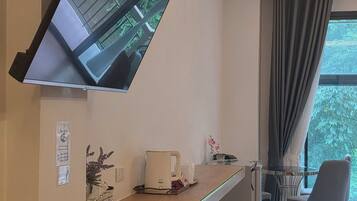 Une chambre d’hôtel moderne dotée d’un téléviseur à écran plat fixé au mur, d’une petite kitchenette, d’un coin salon avec une chaise et une table, et d’une grande fenêtre avec des rideaux.