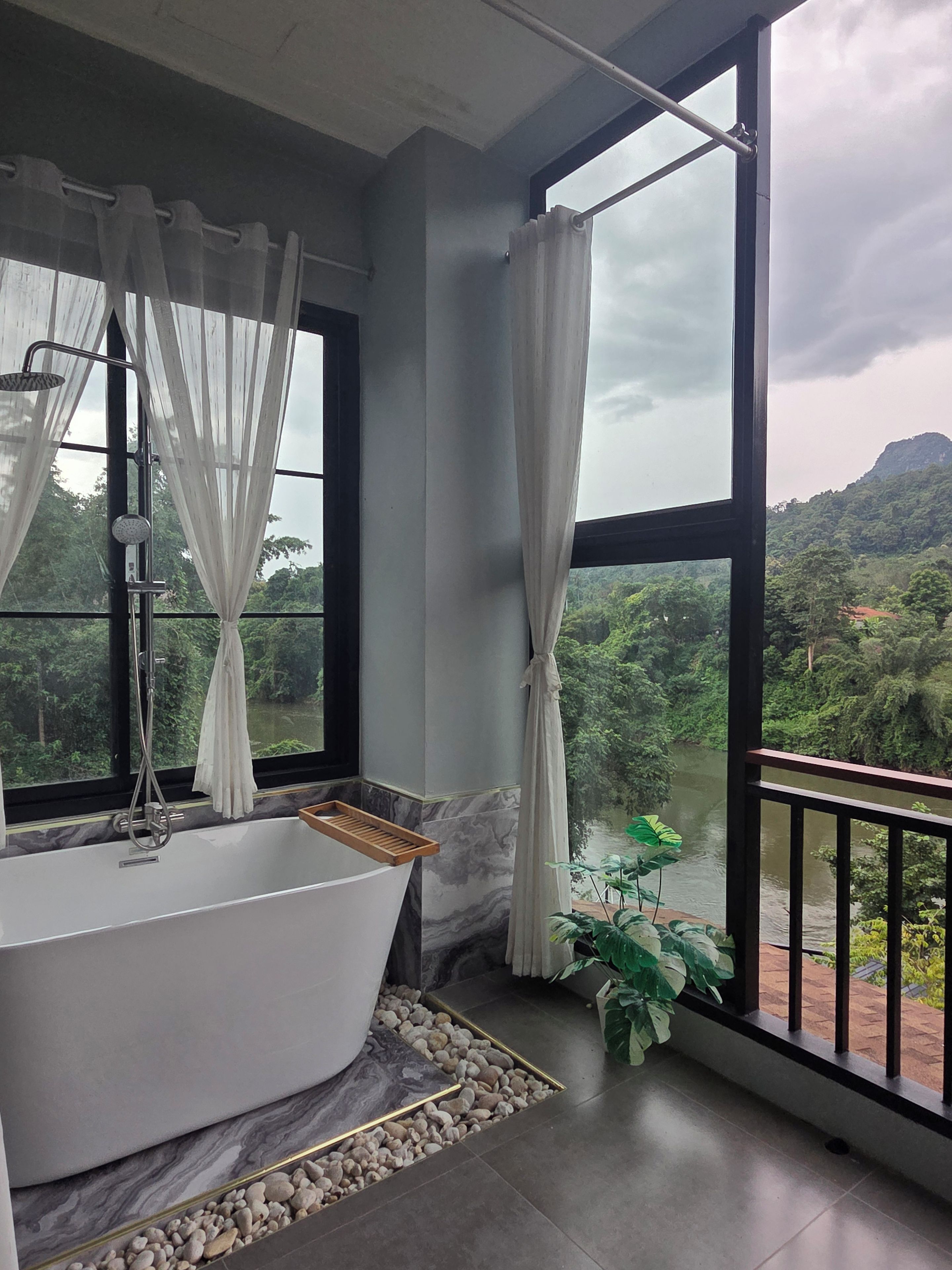 Un baño con una bañera independiente, ducha y vistas a un río y bosque a través de amplios ventanales.