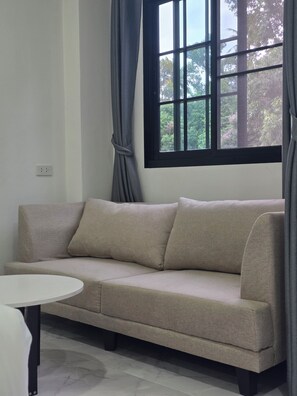 A neutral-toned sofa with a matching armrest, a round white table, and a window with curtains. - KIREERIVER (Thong Pha Phum)