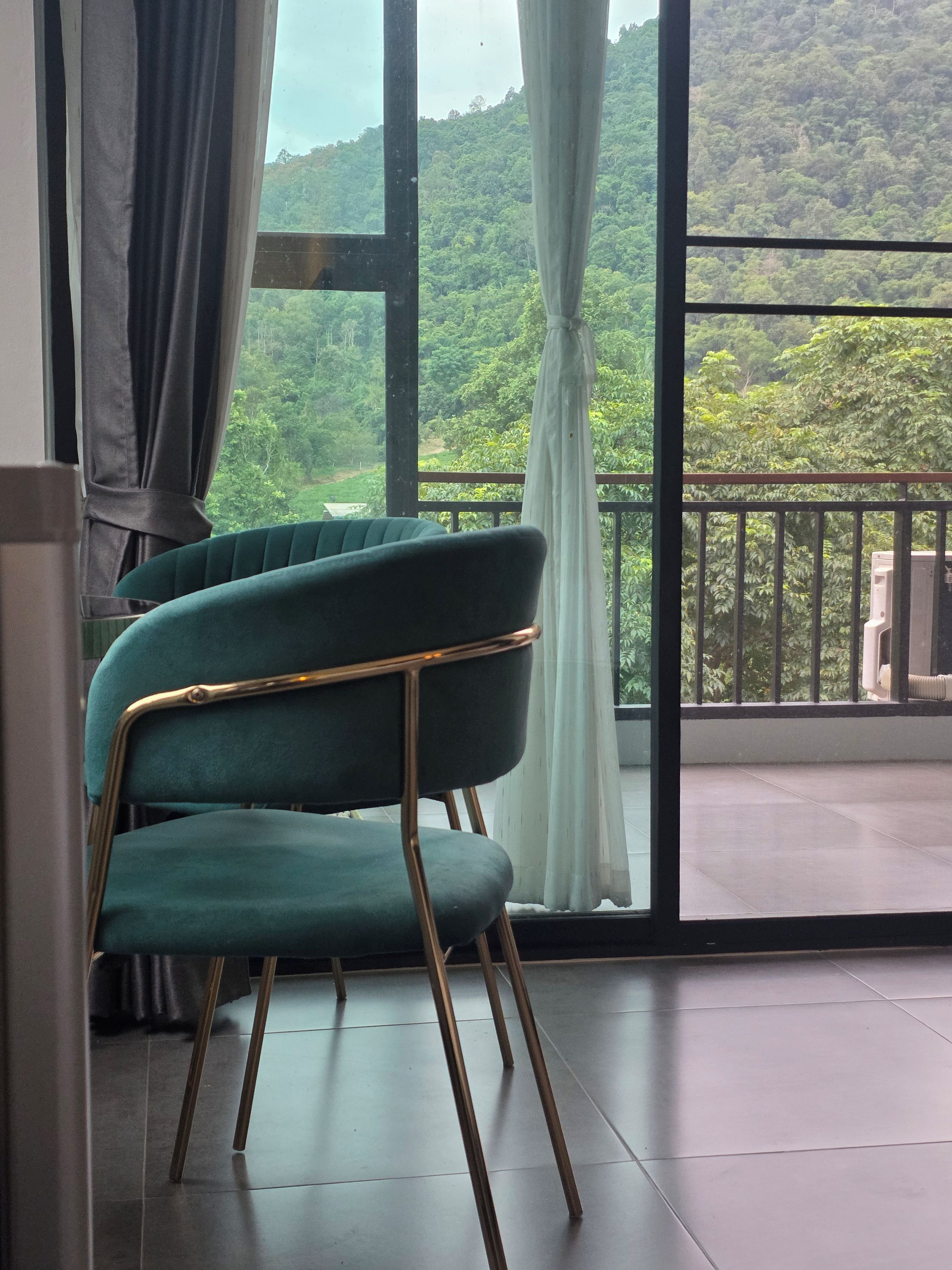Une pièce avec un fauteuil vert, une grande fenêtre avec des rideaux et une vue sur une colline boisée.