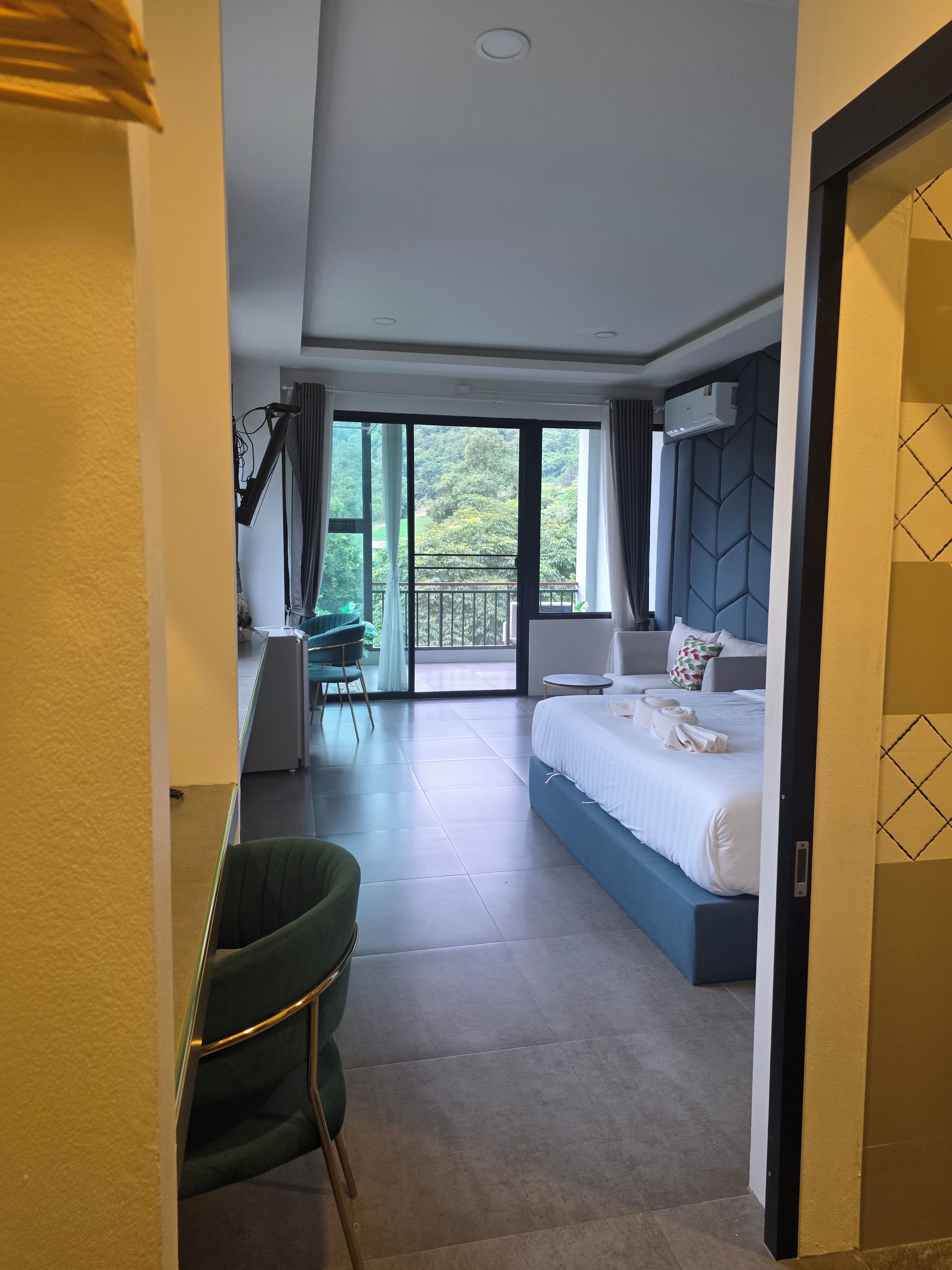 A hotel room with a large bed, a desk, a chair, and a balcony with a view of greenery.
