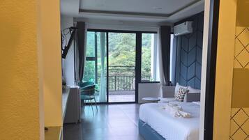 A hotel room with a large bed, a desk, a chair, and a balcony with a view of greenery.