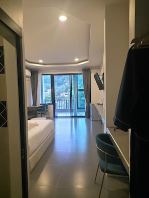 A hotel room with a bed, a desk, a chair, a TV, and a balcony with a view of trees.