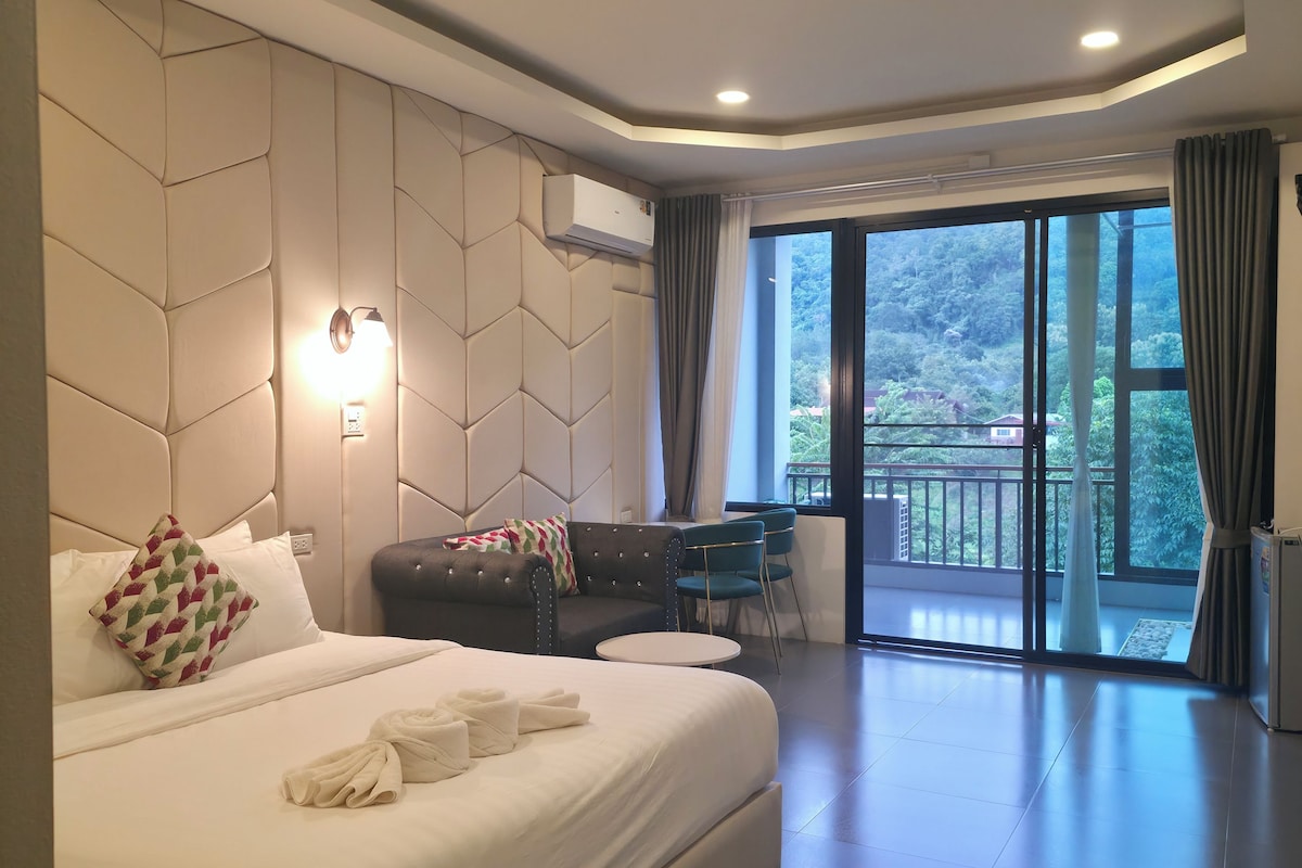 A modern hotel room with a large bed, a seating area with a table, and a balcony with a view.