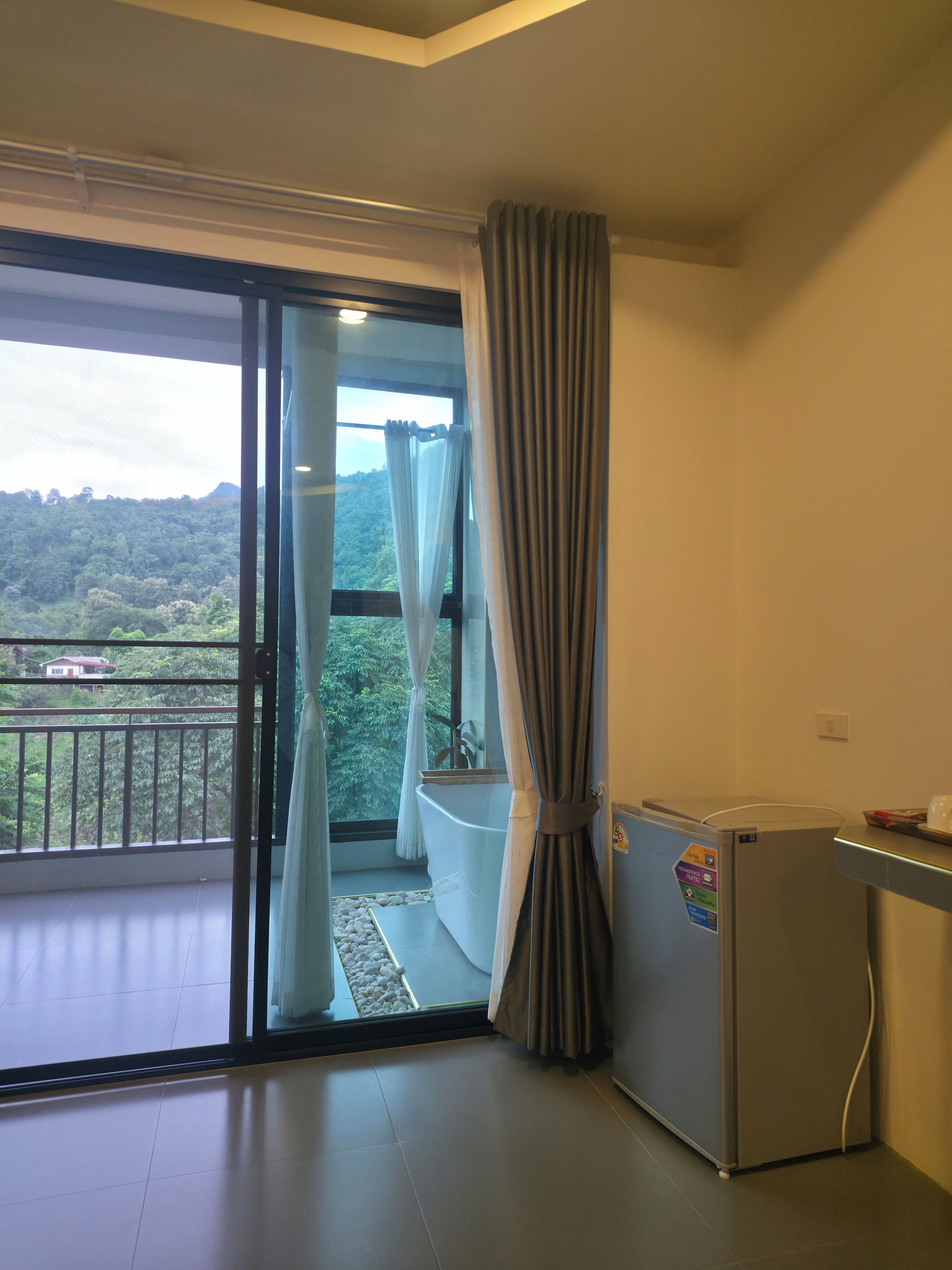 Una habitación con una puerta corrediza de vidrio que da a un balcón, una bañera blanca y un refrigerador pequeño.