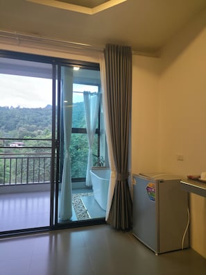 A room with a sliding glass door leading to a balcony, a white bathtub, and a small refrigerator. - KIREERIVER (Thong Pha Phum)