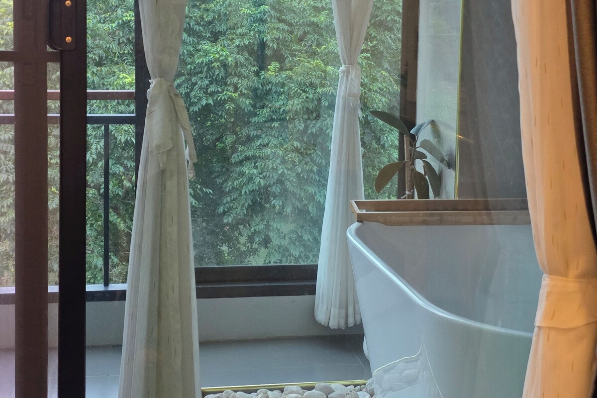 A bathroom with a bathtub, a window with curtains, and a view of a forested area.