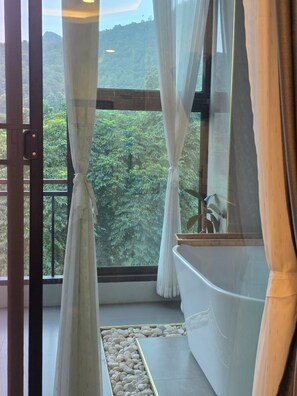 A bathroom with a bathtub, a window with curtains, and a view of a forested area. - KIREERIVER (Thong Pha Phum District)