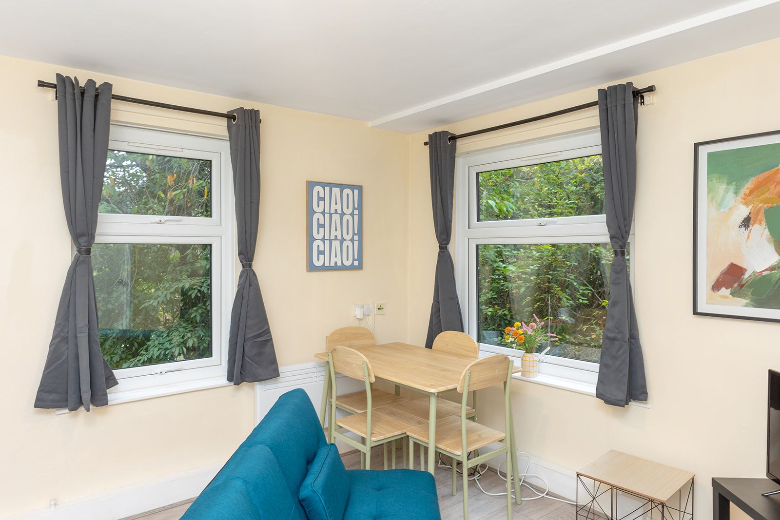 A room with two windows, a table, a chair, a sofa, and a painting on the wall.