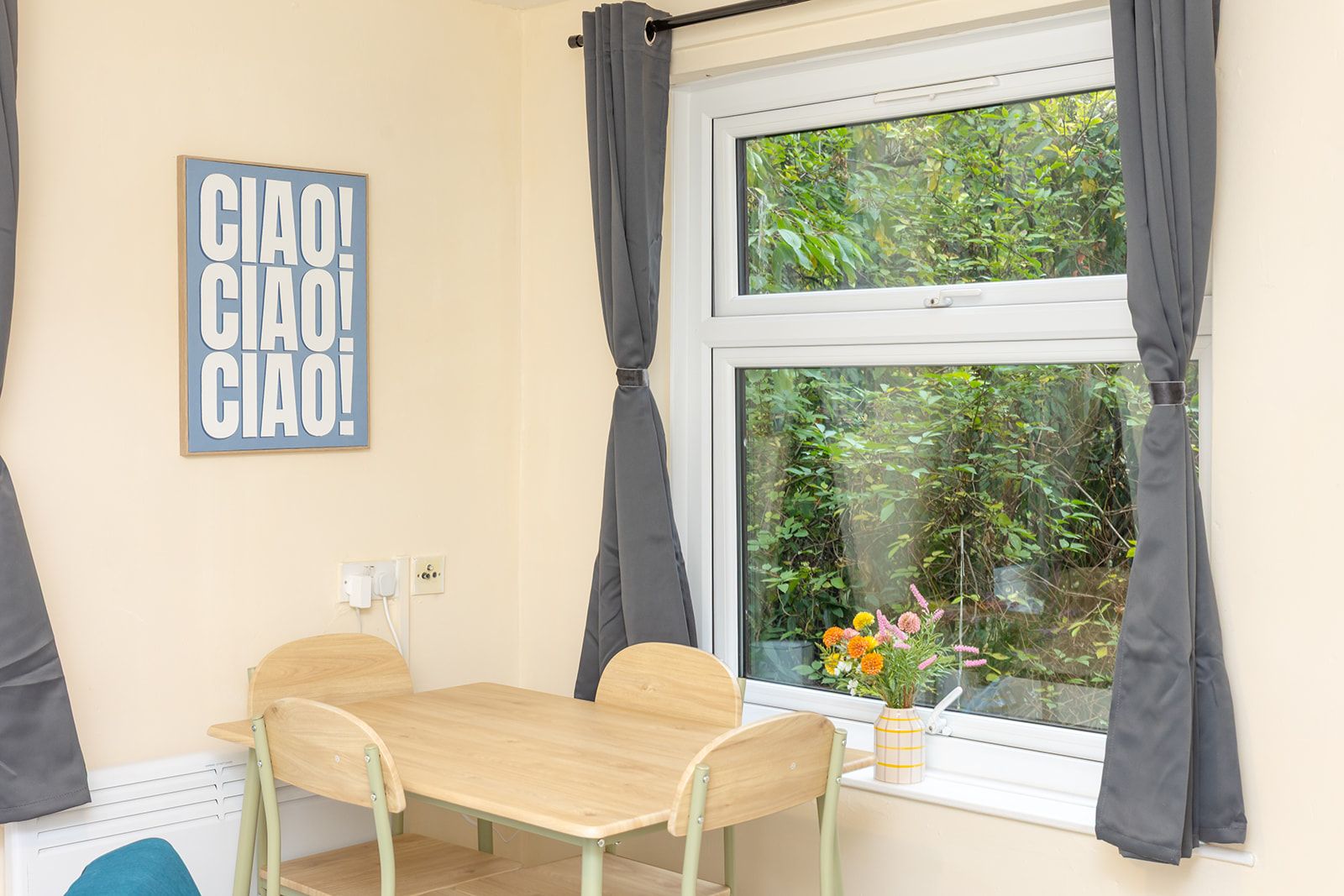 A room with a wooden table, two chairs, a window with curtains, a wall-mounted light switch, and a framed sign.