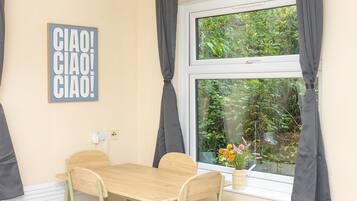 A room with a wooden table, two chairs, a window with curtains, a wall-mounted light switch, and a framed sign.