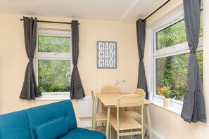 Quarto com um sofá azul, uma mesa de madeira com cadeiras, uma janela com cortinas e uma placa fixada na parede.