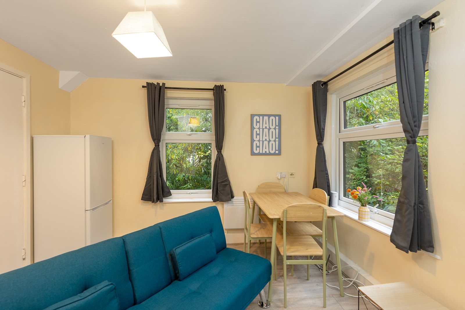 A compact room with a blue sofa, a small dining table with chairs, a white refrigerator, and a window with curtains.