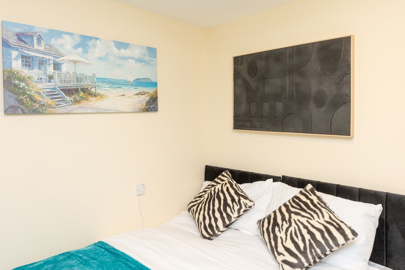 A bedroom with a bed, zebra-patterned pillows, a painting of a beach house, and a geometric abstract artwork.