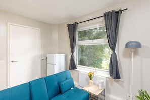 A room with a blue sofa, a white radiator, a white heater, a white wall, a window with grey curtains, and a floor lamp. - 1BR Flat in Stockton | Cozy | Near Station (Stockton-on-Tees)