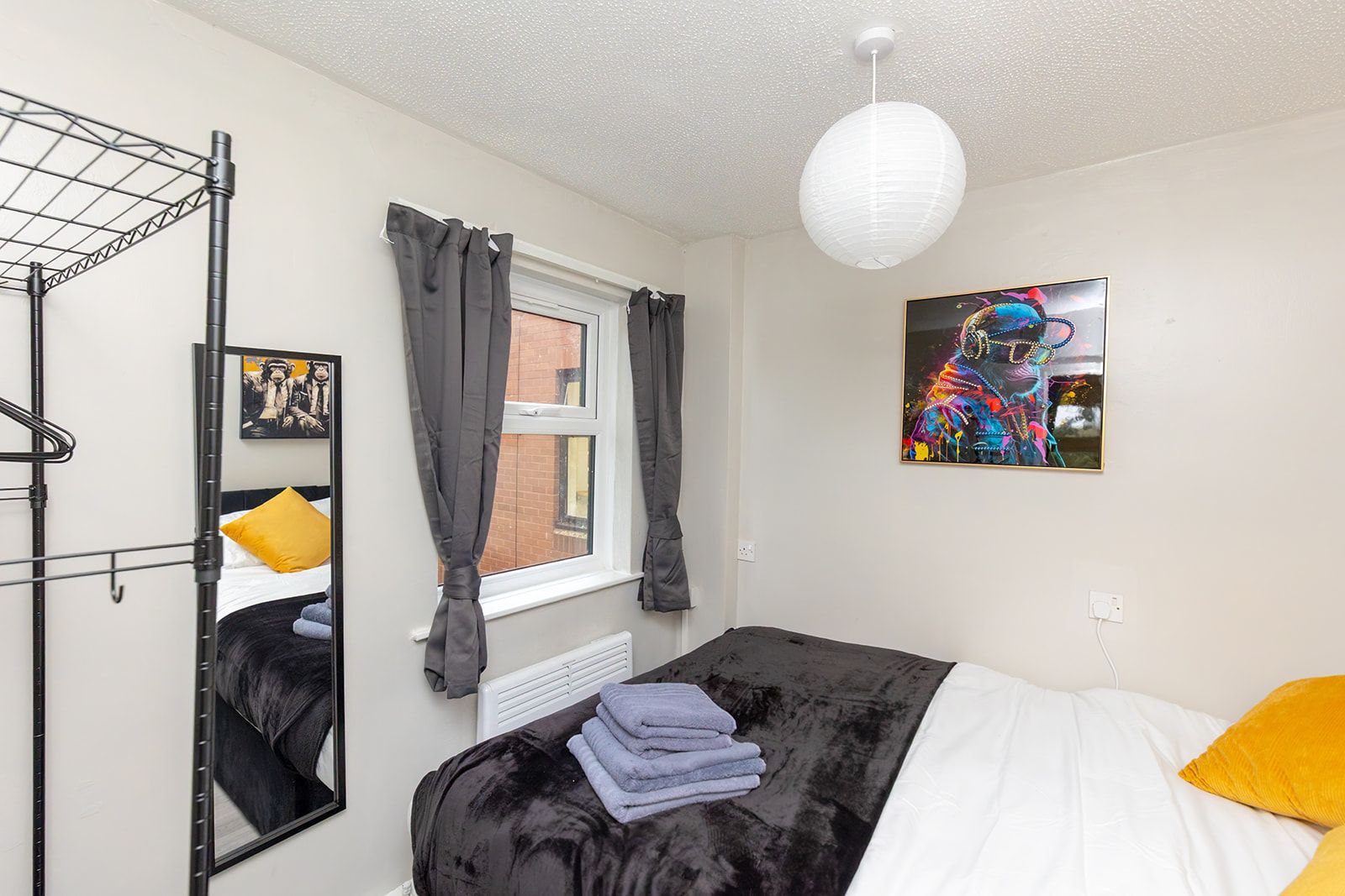 Ein Schlafzimmer mit einem Bett, einem Heizkörper, einer Hängelampe, einem Fenster mit Vorhängen, einem Spiegel und einem an der Wand hängenden Kunstwerk.