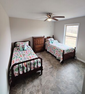 Quarto com duas camas, penteadeira, ventilador de teto e uma janela grande.