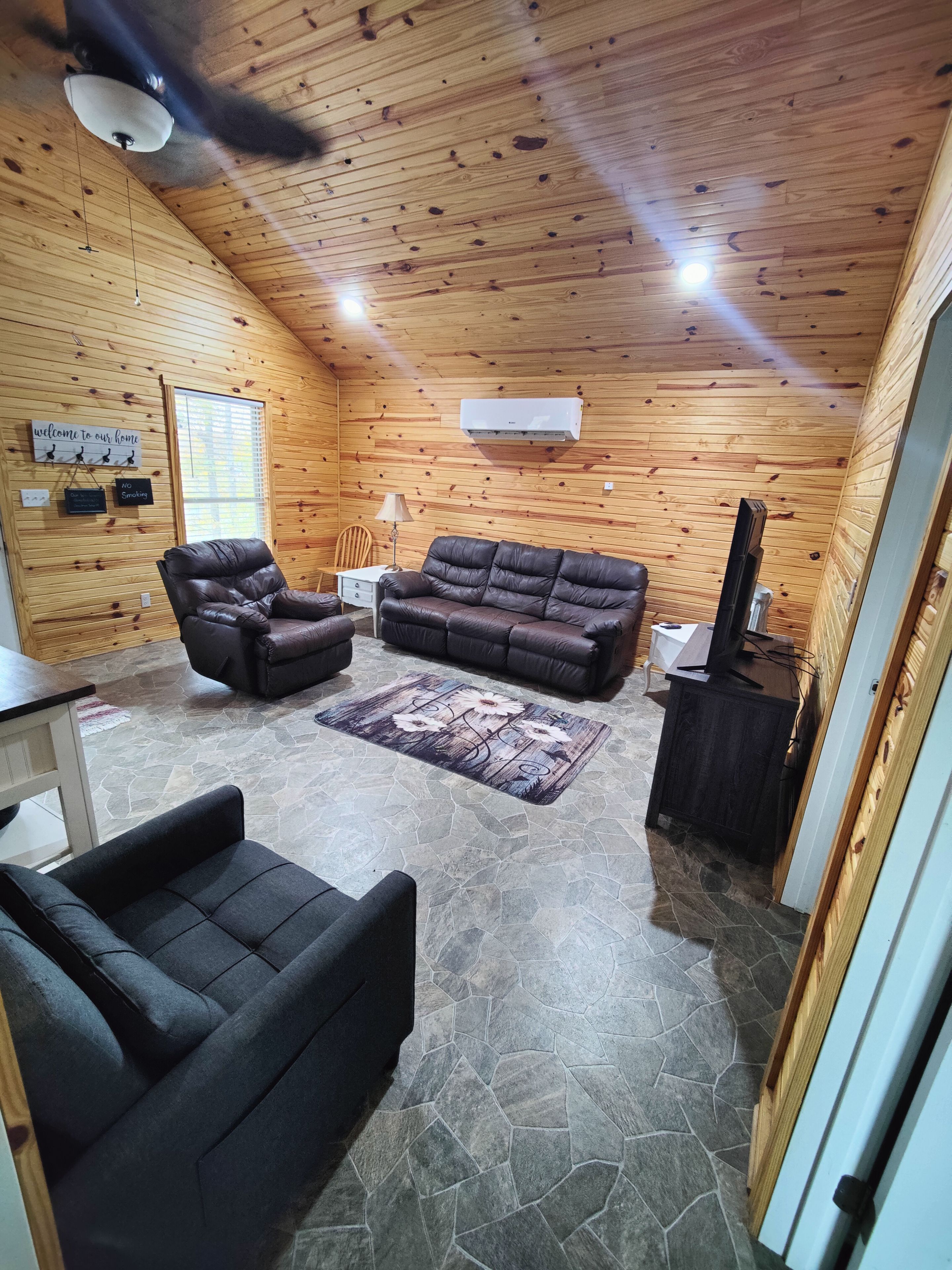 A cozy cabin interior with a stone floor, wooden walls, a leather sofa, a chair, and a television.