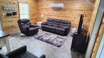 A cozy cabin interior with a stone floor, wooden walls, a leather sofa, a chair, and a television.