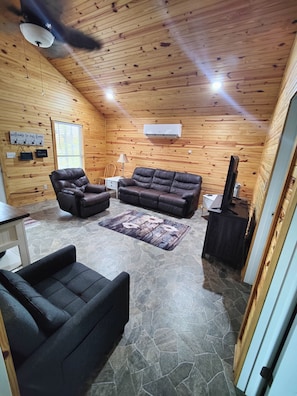 A cozy cabin interior with a stone floor, wooden walls, a leather sofa, a chair, and a television.