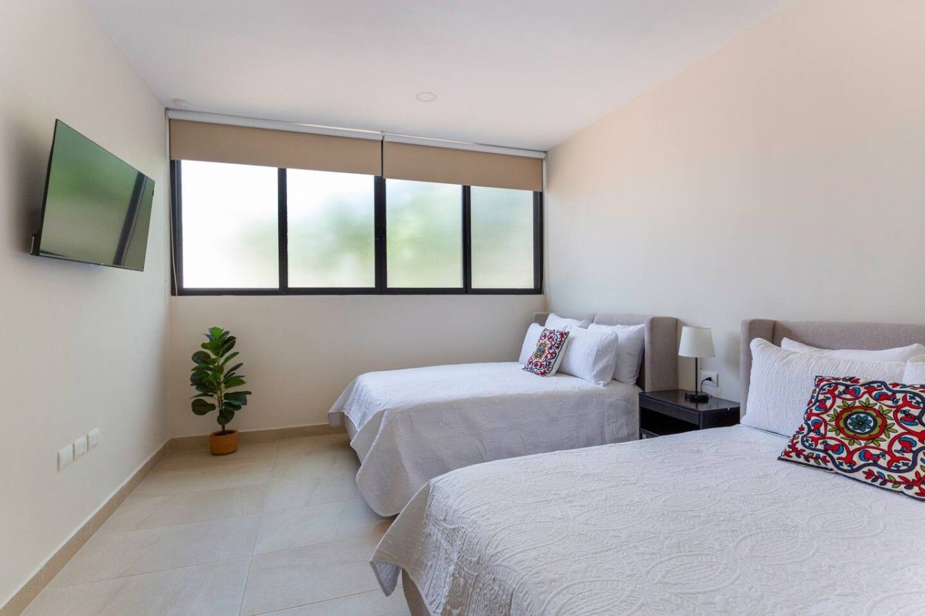 Habitación de hotel con dos camas, un televisor montado en la pared, una planta en maceta y una ventana con persianas.