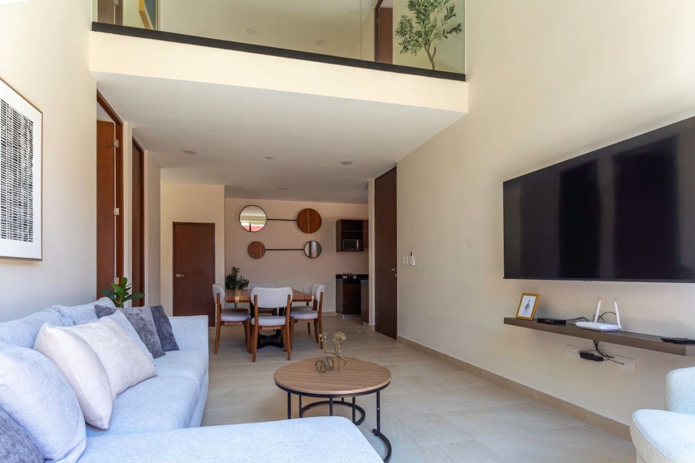 A modern living room with a sofa, a dining table with chairs, a coffee table, a flat-screen TV mounted on the wall, and a staircase leading to an upper level.