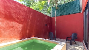 Zona de piscina exterior petita amb dues cadires, envoltada per parets vermelles i una tanca verda.