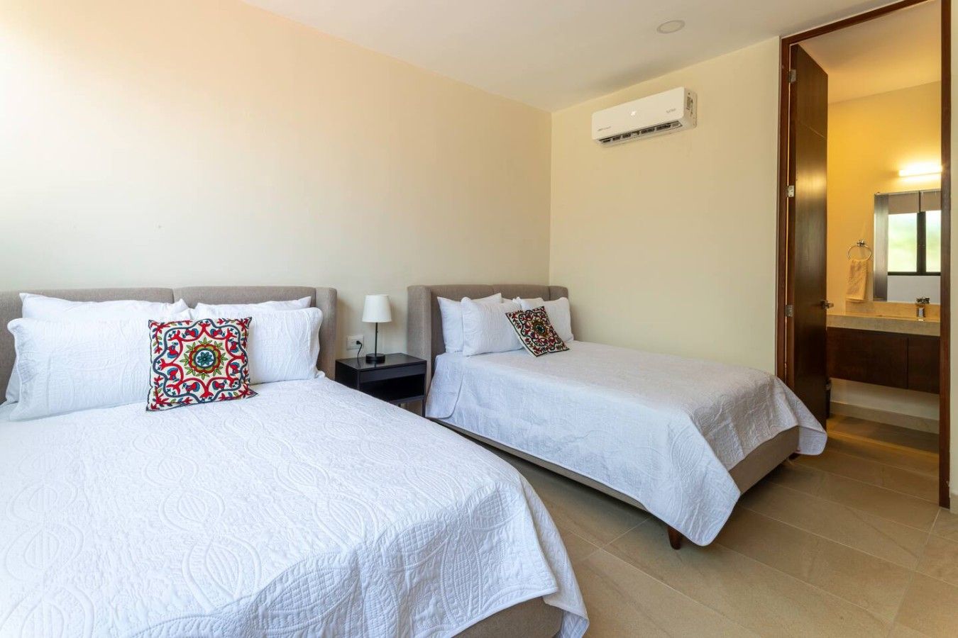 Habitación de hotel con dos camas, una mesita de noche, una lámpara, un equipo de aire acondicionado y un baño con espejo y lavamanos.