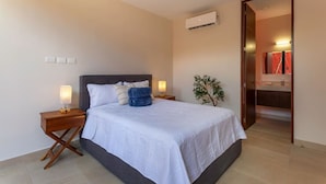 Habitació d'hotel amb llit, taules de nit, làmpada, aparell d'aire condicionat, planta i bany amb mirall i rentatge.