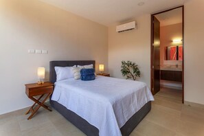 Ein Hotelzimmer mit Bett, Nachttischen, einer Lampe, einer Klimaanlage, einer Pflanze und einem Badezimmer mit Spiegel und Waschbecken.