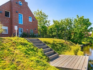 Un bâtiment en briques avec un toit en pente, un escalier en bois menant à une jetée surplombant une étendue d’eau, et une clôture ornée de verdure.