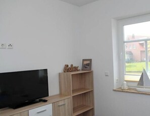 A room with a flat-screen TV on a wooden cabinet, a small shelf with a picture frame and a 'Relax' sign, and a window with a view of a building outside. - Holiday Apartment in Greetsiel With Terrace (Krummhoern)