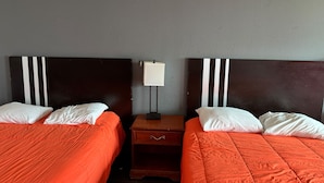 Ein Hotelzimmer mit zwei Betten, jedes mit weißen Kissen und orangefarbener Bettwäsche. Dazwischen befindet sich ein Nachttisch mit einer Lampe.