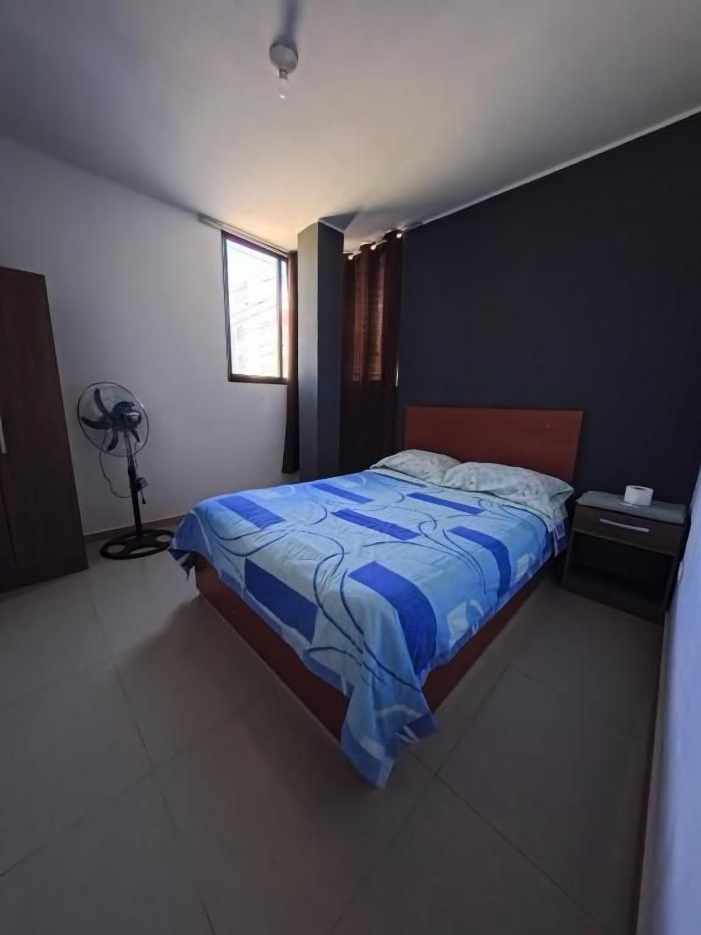 A bedroom with a bed, a bedside table, a nightstand, a fan, and a window.