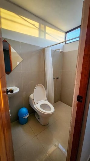 A small bathroom with a toilet, a shower curtain, and a blue trash bin. - VALLE DEL SOL HOTEL (Ica)