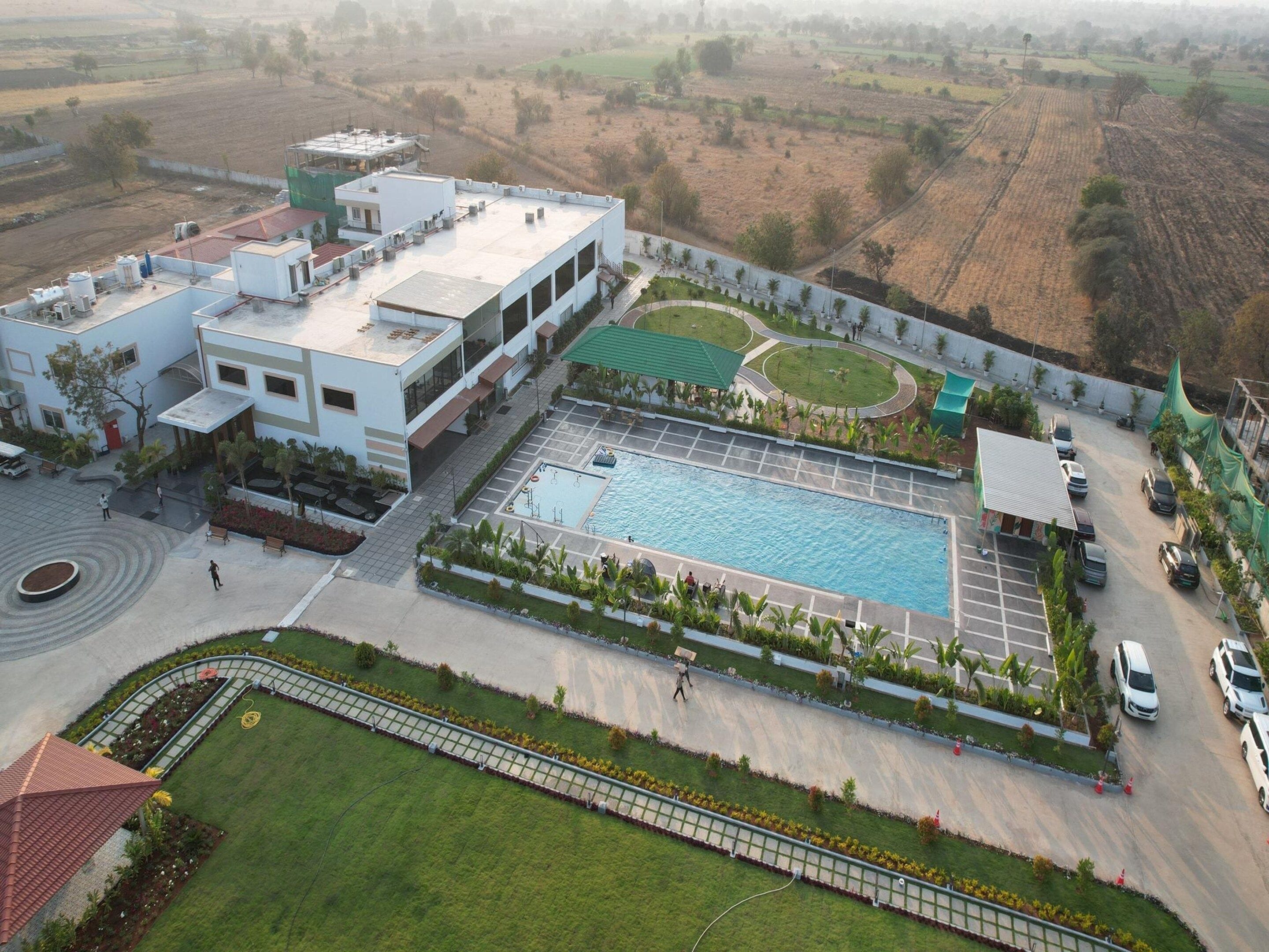 A large building with a swimming pool, surrounded by a landscaped area with a circular garden and a parking lot.