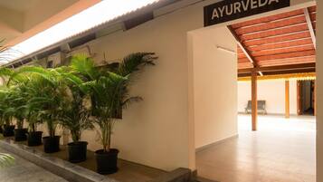 A hallway with potted plants, a sign reading 'AYURVEDA', and a covered area with chairs.