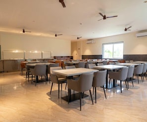 A spacious dining area with multiple tables and chairs, ceiling fans, and a large window providing natural light.
