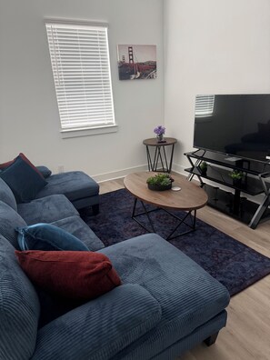 A modern living room with a sectional sofa, a flat-screen TV, a small round table with a plant, and a painting of the Golden Gate Bridge. - Katy Comfort Townhome (Katy)