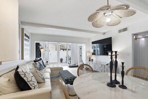 Un salon moderne comprenant un canapé, un pouf, une table à manger et des chaises. Un téléviseur est fixé au mur et un luminaire est suspendu au plafond.
