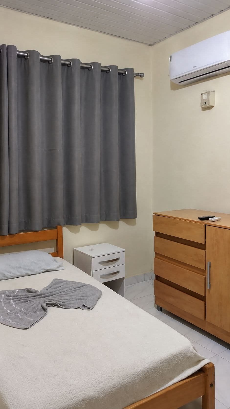 A bedroom with a bed, a dresser, a nightstand, a curtain, and an air conditioning unit.
