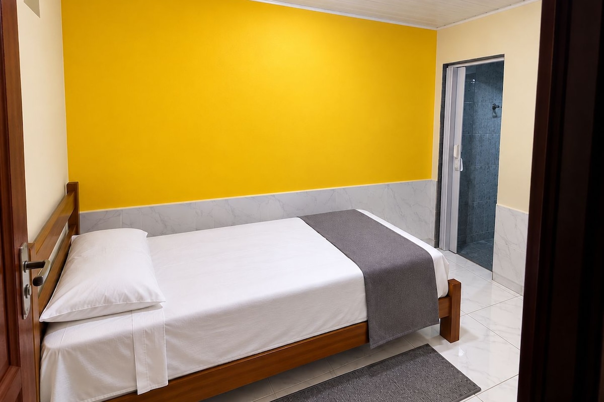 A single bed with a wooden frame, white bedding, and a gray throw blanket in a room with yellow walls and a tiled floor.